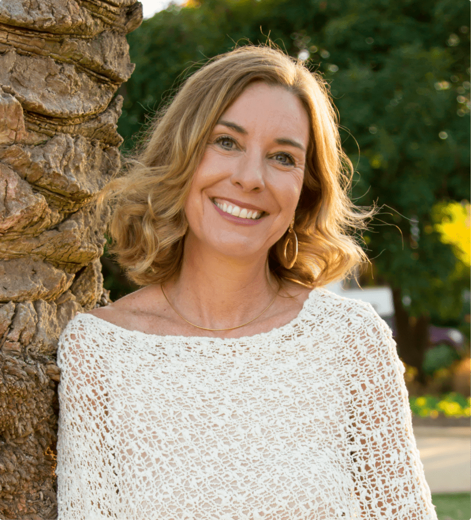 Shawna Robins is leaning against a plam tree wearing a white top and smiling directly at the camera