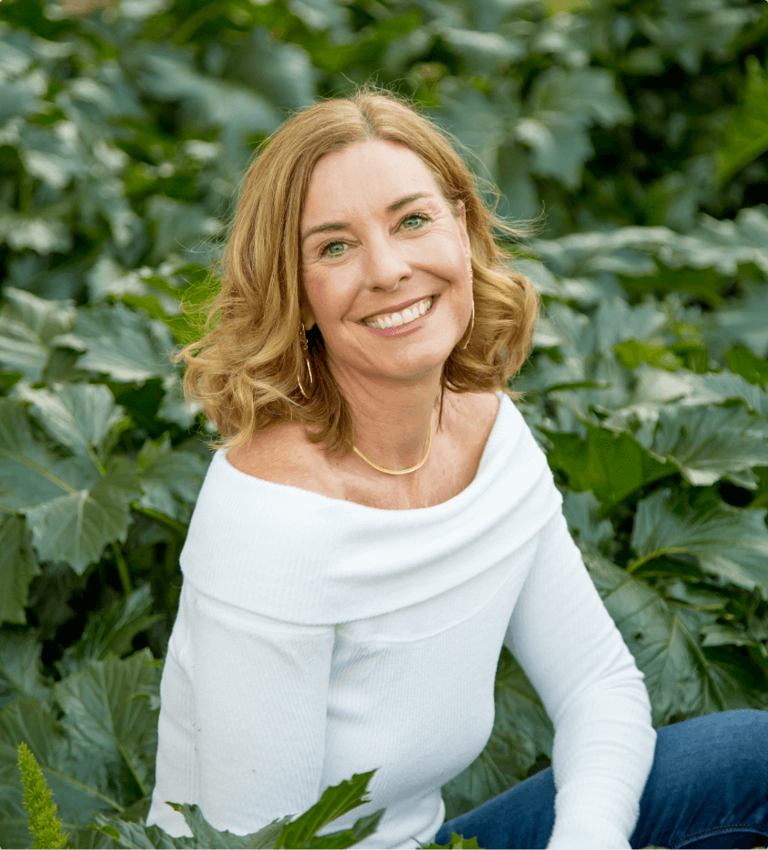 Wellnes coach Shawna Robins crouches in dark cabbage leaves. She is wearing a white off-the-shoulder top and jeans. She has shoulder-length honey-blonde hair and is smiling at the camera.